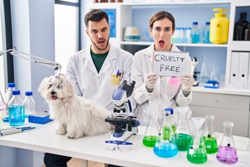 Young hispanic people working at scientist laboratory with dog in shock face, looking skeptical and sarcastic, surprised with open mouth