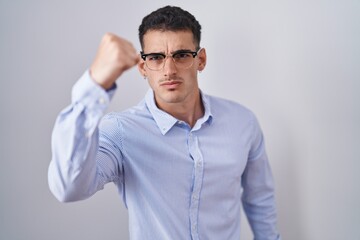 Handsome hispanic man wearing business clothes and glasses angry and mad raising fist frustrated and furious while shouting with anger. rage and aggressive concept.