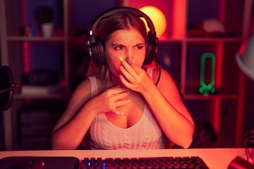 Young blonde woman playing video games wearing headphones smelling something stinky and disgusting, intolerable smell, holding breath with fingers on nose. bad smell - obrazy, fototapety, plakaty