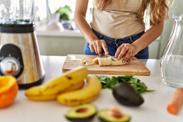 Young beautiful hispanic woman preparing vegetable smoothie with blender cutting banana at the kitchen