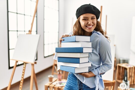 Young beautiful hispanic woman artist smiling confident holding books at art studio