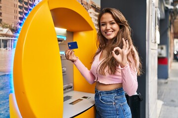 Young hispanic woman taking cash from atm with credit card doing ok sign with fingers, smiling...