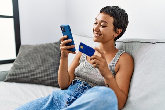 Young hispanic woman using smartphone holding credit card at home