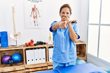 Middle age hispanic physiotherapist woman working at pain recovery clinic laughing at you, pointing finger to the camera with hand over mouth, shame expression