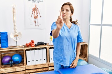 Middle age hispanic physiotherapist woman working at pain recovery clinic pointing with finger up and angry expression, showing no gesture