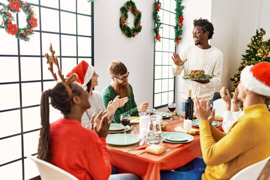Group of people meeting clapping and sitting on the table. Man standing and holding roasted turkey celebrating Christmas at home.