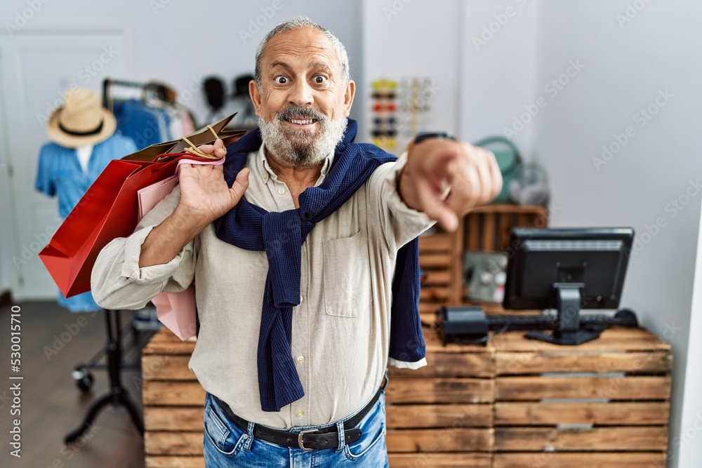 Poster handsome senior man holding shopping bags at boutique shop pointing to you and the camera with finge