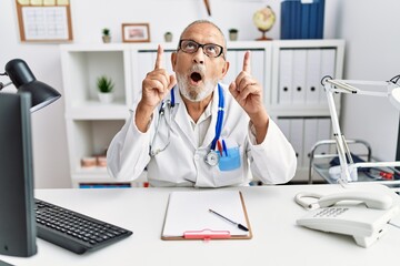 Obraz premium Mature doctor man at the clinic amazed and surprised looking up and pointing with fingers and raised arms.