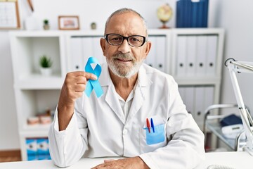 Mature doctor man holding blue ribbon at the clinic looking positive and happy standing and smiling...