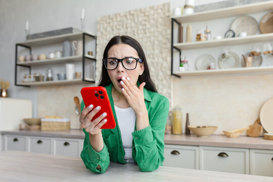 A young woman looks shocked and surprised at the phone, covered her mouth with her hand, saw a message from her ex-lover, learned about betrayal. Sitting at home in the kitchen and holding a red phone