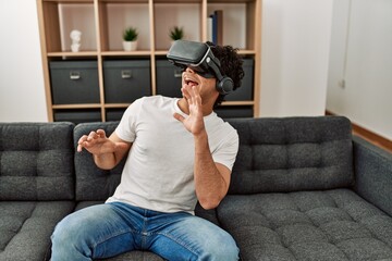Obraz premium Young hispanic man scared playing with virtual reality glasses sitting on the sofa at home.