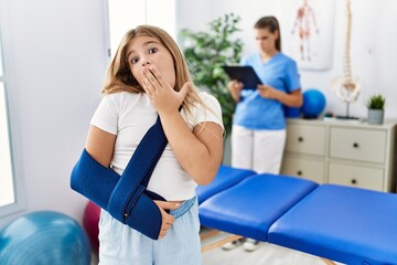 Blonde little girl wearing arm on sling at rehabilitation clinic covering mouth with hand, shocked...