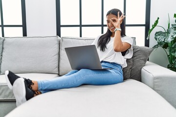 Fototapeta premium Young hispanic woman using laptop at home sitting on the sofa smiling happy doing ok sign with hand on eye looking through fingers