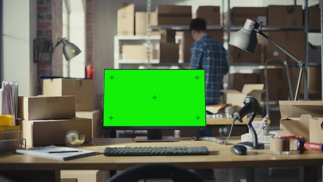 Desktop Computer Monitor Standing on a Table with a Green Screen Chromakey Mock Up Display. Small Business Storage Room with Worker Walking in the Background. Desk with Cardboard Boxes. Static Shot.