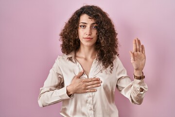 Hispanic woman with curly hair standing over pink background swearing with hand on chest and open...