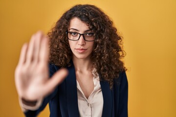Hispanic woman with curly hair standing over yellow background doing stop sing with palm of the hand. warning expression with negative and serious gesture on the face.