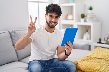 Hispanic man with beard using touchpad sitting on the sofa smiling with happy face winking at the...