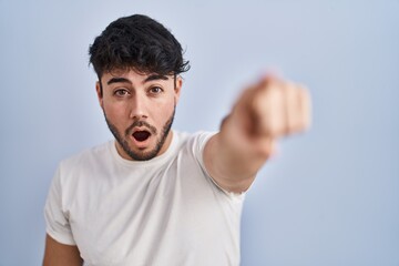Hispanic man with beard standing over white background pointing with finger surprised ahead, open mouth amazed expression, something on the front