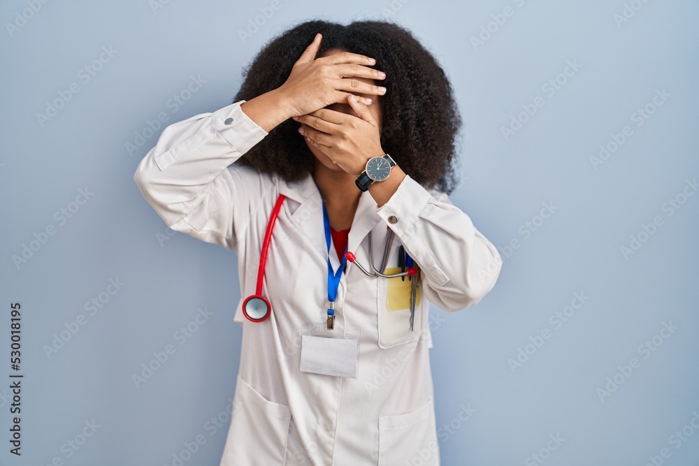 Poster Young african american woman wearing doctor uniform and stethoscope covering eyes and mouth with hands, surprised and shocked. hiding emotion