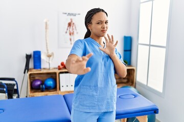 Young african american woman working at pain recovery clinic disgusted expression, displeased and fearful doing disgust face because aversion reaction. with hands raised