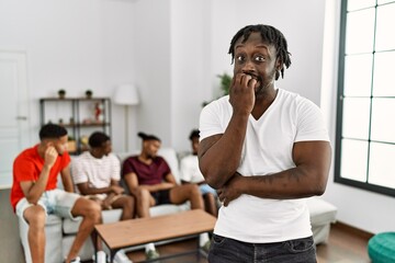 Young african man with friends at the living room looking stressed and nervous with hands on mouth biting nails. anxiety problem.