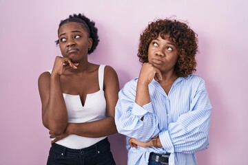 Two african women standing over pink background with hand on chin thinking about question, pensive expression. smiling with thoughtful face. doubt concept.