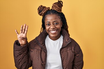 African woman with braided hair standing over yellow background showing and pointing up with fingers number four while smiling confident and happy.