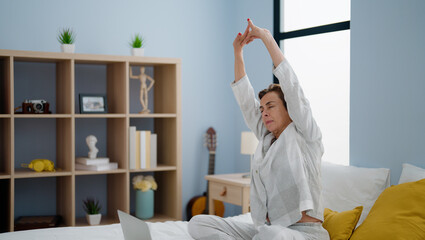 Middle age hispanic woman having online yoga session sitting on bed at bedroom