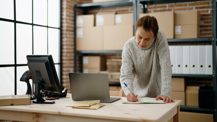 Middle age hispanic woman ecommerce business worker writing on book using laptop at storehouse...