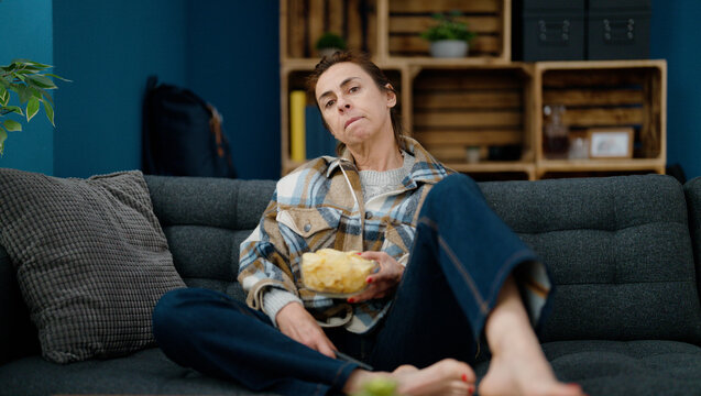 Middle Age Hispanic Woman Eating Chips Potatoes Watching Movie At Home