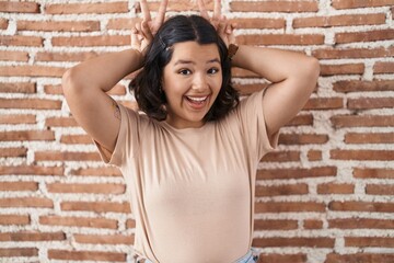 Young hispanic woman standing over bricks wall posing funny and crazy with fingers on head as bunny ears, smiling cheerful