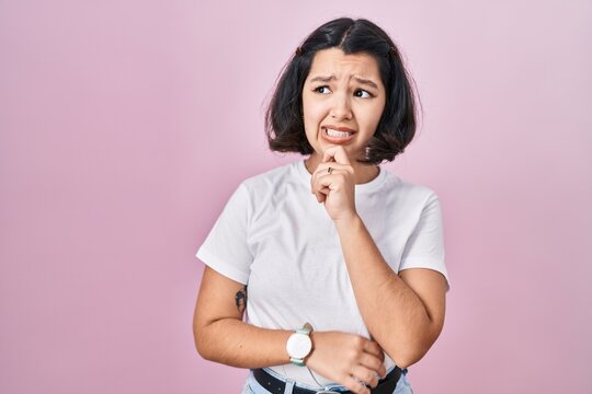 Young hispanic woman wearing casual white t shirt over pink background thinking worried about a question, concerned and nervous with hand on chin