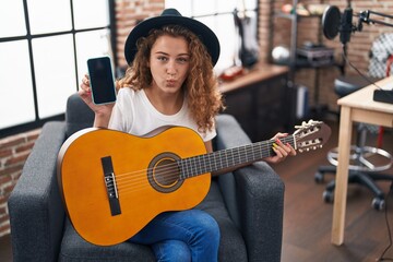 Young caucasian woman playing classic guitar at music studio holding smartphone puffing cheeks with funny face. mouth inflated with air, catching air. - obrazy, fototapety, plakaty