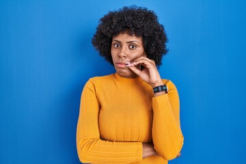 Black woman with curly hair standing over blue background mouth and lips shut as zip with fingers....