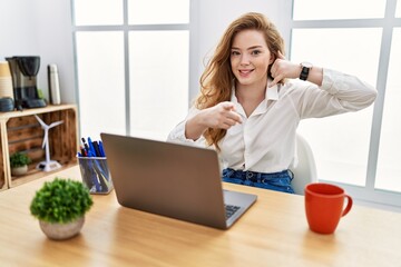 Young caucasian woman working at the office using computer laptop smiling doing talking on the telephone gesture and pointing to you. call me.