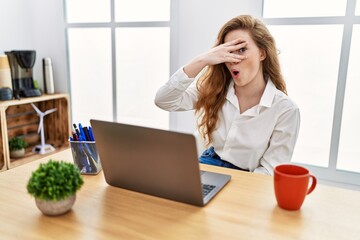 Obraz premium Young caucasian woman working at the office using computer laptop peeking in shock covering face and eyes with hand, looking through fingers with embarrassed expression.
