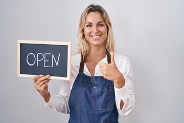 Young blonde woman wearing apron holding blackboard with open word smiling happy and positive, thumb up doing excellent and approval sign