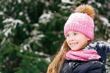 Little girl in pink hat is looking aside with a smile in winter forest.