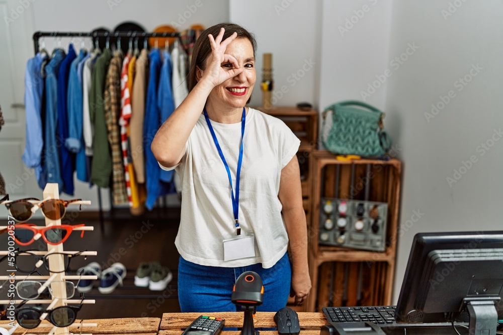 Wall mural Beautiful middle age hispanic woman working as manager at retail boutique doing ok gesture with hand smiling, eye looking through fingers with happy face.