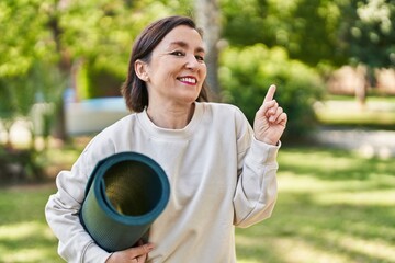 Middle age hispanic woman holding yoga mat at the park smiling happy pointing with hand and finger to the side