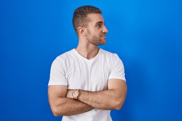 Young caucasian man standing over blue background looking to the side with arms crossed convinced and confident