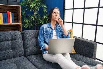 Hispanic woman using laptop at home serious face thinking about question with hand on chin,...