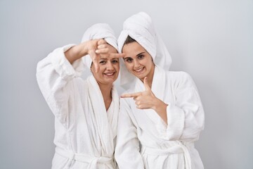 Middle age woman and daughter wearing white bathrobe and towel smiling making frame with hands and fingers with happy face. creativity and photography concept.