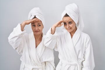 Middle age woman and daughter wearing white bathrobe and towel very happy and smiling looking far away with hand over head. searching concept.