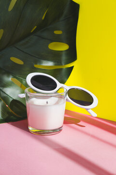 Sunglasses in the style of the 90s with a white frame on a white candle in a glass on a tropical background.