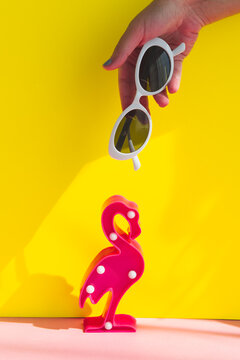 A hand holds trendy stylish sunglasses in a white frame over a lamp in the shape of a pink flamingo. A conceptual idea.