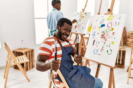 African american painter couple smiling happy painting at art studio. Man holding paintbrushes sitting on chair.