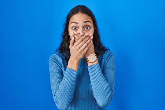 Young brazilian woman standing over blue isolated background shocked covering mouth with hands for mistake. secret concept.
