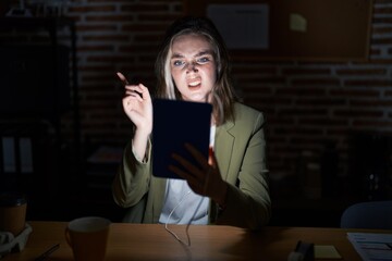 Blonde caucasian woman working at the office at night pointing aside worried and nervous with forefinger, concerned and surprised expression