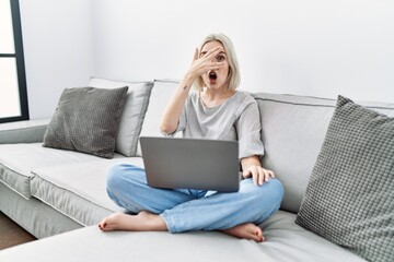 Fototapeta premium Young caucasian woman using laptop at home sitting on the sofa peeking in shock covering face and eyes with hand, looking through fingers with embarrassed expression.